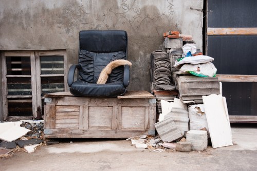 A black office chair with a fabric covering and curved armrests is positioned on a wooden pallet. To the right, there are stacks of construction materials, including cardboard boxes, plastic-wrapped items, and concrete blocks, some of which are leaning or piled unevenly on the ground. The background features a weathered, beige wall with visible dirt and grime, and a small vent with horizontal slats near the bottom left. The scene appears to be outdoors, possibly on a driveway or service area, with scattered debris such as broken pieces of board and small stones nearby. This setup suggests a location involved in rubbish or renovation waste, which a professional rubbish removal service like Garden Clearance Norbiton could help clear from the premises in the Norbiton area, near Kingston or southwest London.