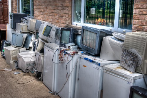 A row of discarded obsolete computer monitors and electronic devices, including CRT and flat-screen screens, arranged outdoors on a concrete pavement next to a brick wall with large windows reflecting trees and greenery. The monitors vary in size, color, and condition, with some showing signs of damage or disassembly, and tangled wires are visible among the stacked equipment. There are no visible packaging materials or other waste items; the collection appears to be a pile of e-waste awaiting disposal or recycling. The background features a brick building façade with a window, indicating an urban or residential setting, possibly in Norbiton or nearby areas. The scene demonstrates a typical e-waste accumulation that garden clearance services like those offered by Garden Clearance Norbiton may assist in removing from garden or property clearance projects, highlighting the importance of proper electronic waste disposal.