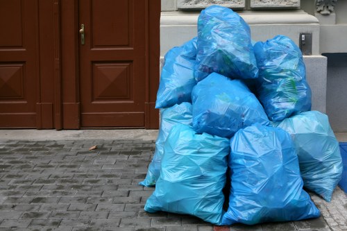 A collection of multiple large blue plastic rubbish bags arranged in a pile on a paved pavement in front of a brown wooden door. The bags appear full and are tightly knotted at the top, with some showing slight creases and folds in the plastic material. The background includes the lower part of a cream-colored building's wall and a grey utility box or pipe on the right side of the image. The scene suggests a rubbish removal collection outside a residential property in Norbiton, with the bags ready for disposal or collection by a local waste management service such as Garden Clearance Norbiton. The area is clean, and the bags occupy the foreground, with the paving providing a textured surface beneath them, highlighting the typical outdoor environment for garden or household waste disposal activities.