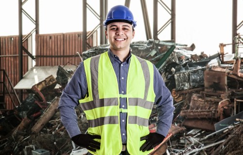 A young male waste management worker wearing a blue safety helmet, high-visibility yellow vest, black gloves, and a blue shirt stands with his hands on his hips in an indoor recycling or waste storage facility. Behind him, there is a large pile of assorted scrap metal, including various steel and iron objects with different textures and finishes, some rusted and others painted. The environment features a metal framework structure and natural light coming from upper windows, illuminating the scene. The image conveys a professional scene related to rubbish removal services, with visual cues suggesting a focus on industrial or garden clearance in Norbiton, possibly associated with Garden Clearance Norbiton. The overall setting emphasizes the handling of bulky, metallic waste in a controlled environment, supporting local rubbish management expertise.
